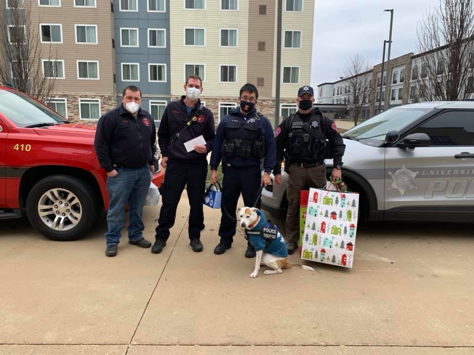 A dog and police officers holding gift bags pose for a photo in front of an apartment complex.