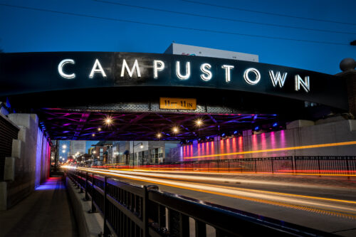 Long-exposure image of Campustown sign on viaduct on Green Street as light is captured from vehicles moving along the street. Businesses are visible along the street in the background.