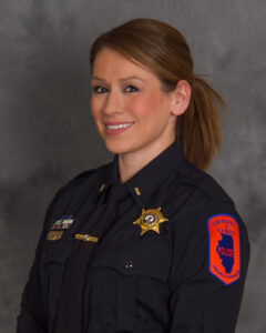 Smiling female police officer in a dark uniform, with badges on chest, against a gray backdrop.