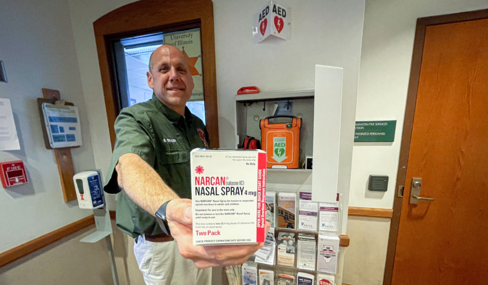 Brian Brauer standing next to an AED and displaying a box of Narcan.