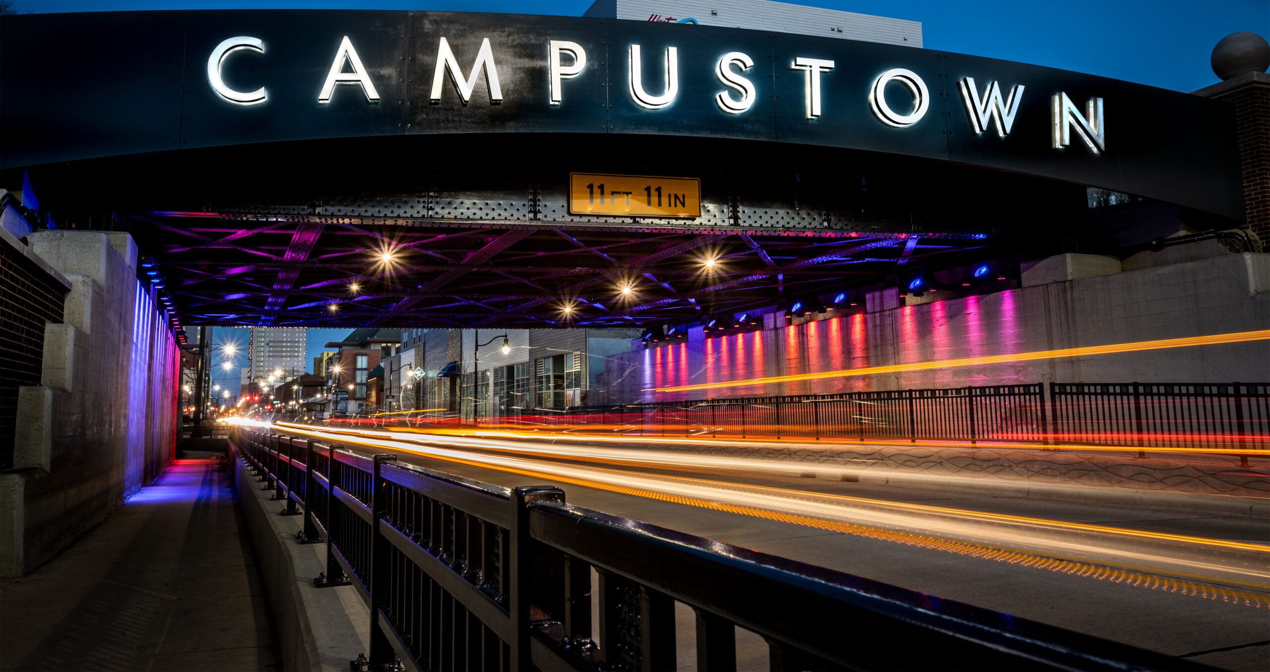 Campustown sign on viaduct over Green Street with time-lapse images of vehicle lights streaming underneath it.