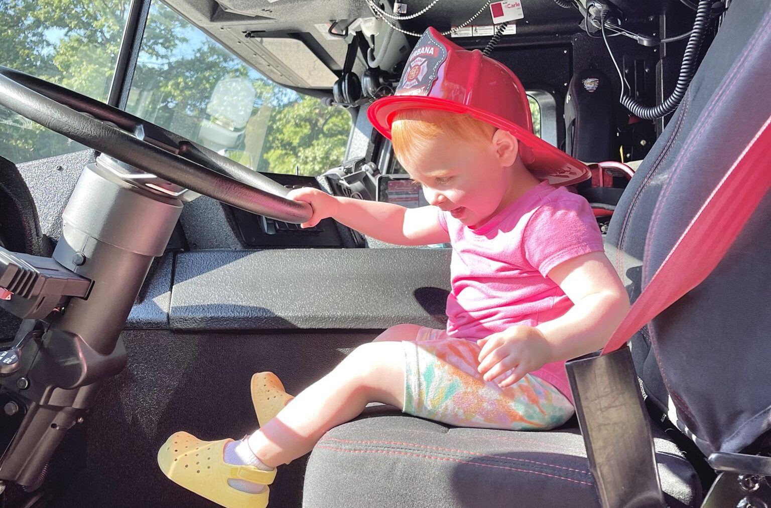 A young girl in a plastic fire department toy helmet sits at the wheel of a fire truck.